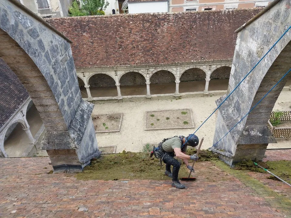 Cordiste retirant les mousses à la main sur la toiture en tuiles de l’église de Donnemarie-Dontilly.